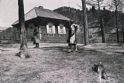 Two individuals in traditional alpine attire stand near a rustic wooden cabin with a peaked roof, framed by a simple metal fe...