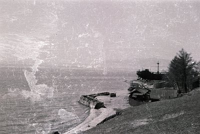 Vintage black-and-white coastal scene showing abandoned concrete structures and a partially submerged shipwreck on rocky shor...