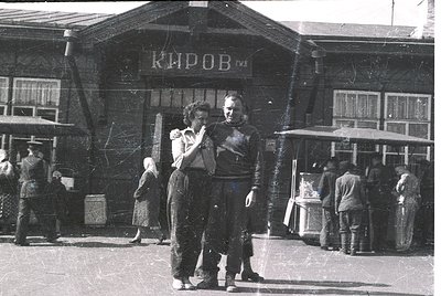 Couple dancing near a Soviet-era "Киров" (Kirov) building, likely a train station or cultural center, in mid-20th century. Wo...