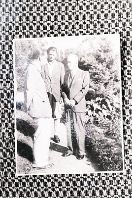 Three men in formal 1950s–60s attire pose outdoors near lush greenery. The central figure wears a suit with a pocket square, ...
