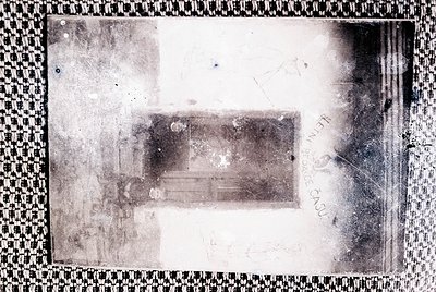 Vintage sepia-toned negative showing a blurred interior with faint furniture—likely a bed or sofa—against a wall. Checkered b...