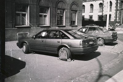 1970s-era sedan parked on cobblestone street with Soviet-style parking block. Neo-classical building with arched windows and ...