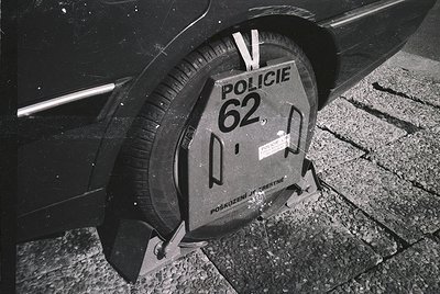 Black-and-white close-up of a Polish police wheel clamp labeled "Policja 62" on a vehicle tire, indicating a parking violatio...