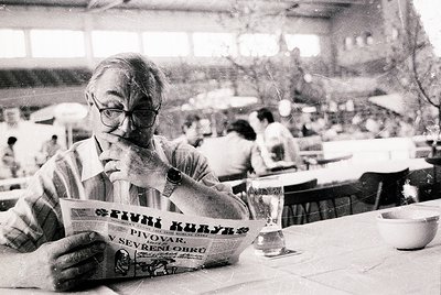 Mid-20th century café scene: Man in striped shirt reads *Pivni Kuliyar* beer menu (Bulgarian: "Beer Tavern") at a seaside or ...