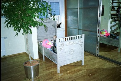 Vintage wooden crib (1928) with "Jessie" engraved, positioned under a potted plant in a dimly lit room. Wooden floor and meta...