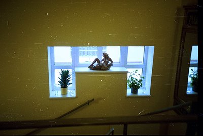 A person sits on a narrow window sill in a dimly lit interior, framed by white-painted casement windows. Two potted plants fl...