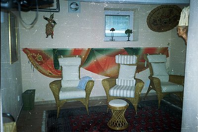 Vintage indoor seating area with mid-century modern wicker chairs, striped cushions, and a woven stool. Bold geometric wallpa...
