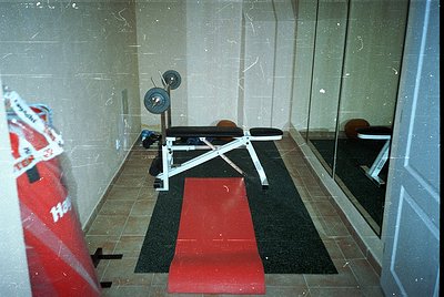 Vintage gym bench press station in a tiled, utilitarian space. White adjustable bench with weight stack, red rubber mat, and ...