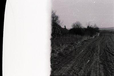 Vintage black-and-white rural roadside scene with plowed field and sparse vegetation. Dusty track curves through open landsca...