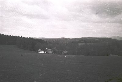 Vintage black-and-white rural landscape featuring a single-story farmhouse nestled in a forested valley. Open fields dominate...