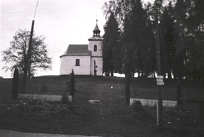 Small white church with a single bell tower and cross-topped dome, perched on a raised grassy mound. Surrounded by a low meta...