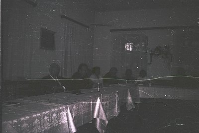 Vintage black-and-white photo of a dimly lit indoor gathering, likely a 1960s–1970s event. Round table draped in ornate table...
