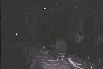Vintage black-and-white shot of a dimly lit indoor dining scene, likely mid-20th century. Table set with white tablecloth, cr...