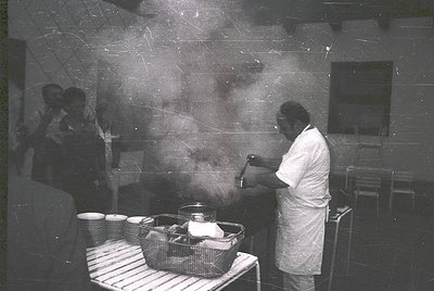 Black-and-white industrial kitchen scene from the 1960s–70s. Chef in white uniform ladles steaming liquid from a pot, surroun...