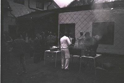 Vintage black-and-white photo of a small, dimly lit courtyard gathering, likely 1960s–1970s. A man in a white shirt and rolle...