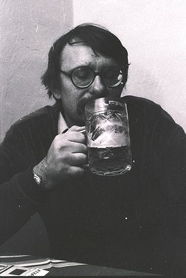 Mid-20th century man in round glasses sips from a frosted beer mug, likely in a café or pub setting. His attire suggests a fo...