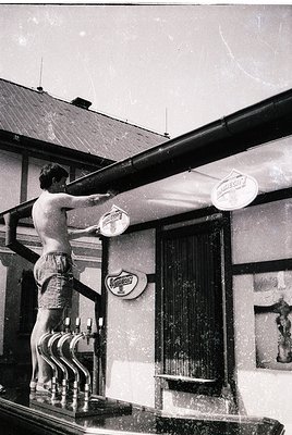 Mid-20th century black-and-white shot of a man in swim trunks washing a neon beer sign on a balcony railing. Wooden building ...