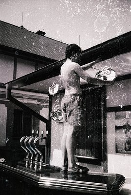 Vintage black-and-white photo of a man in swimwear cleaning a brass bar rail in a 1950s-60s American-style bar. Industrial-er...