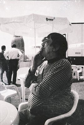 Mid-century outdoor café scene featuring a man in a striped sweater smoking, seated at a white plastic chair. Umbrellas and v...