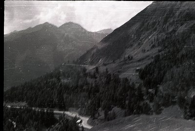 Vintage black-and-white aerial shot of a winding mountain road flanked by dense coniferous forest, leading toward jagged peak...