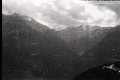 Vintage black-and-white aerial view of rugged alpine terrain with dense cloud cover. Visible grid lines suggest a topographic...