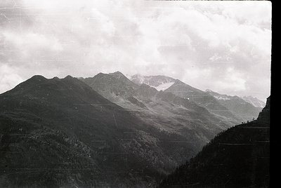 Black-and-white alpine landscape featuring jagged peaks and dense forest valleys, likely European Alps. Snow patches highligh...