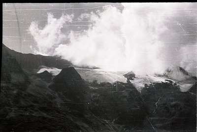 Volcanic eruption plume billowing over rugged terrain, likely a stratovolcano. Dense ash cloud obscures distant slopes, sugge...