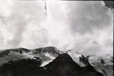 Vintage black-and-white aerial view of rugged volcanic terrain with jagged peaks and steep slopes. Dense cloud cover obscures...