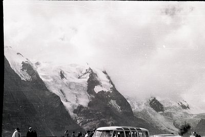 Snow-capped alpine peaks dominate this vintage black-and-white photo, likely from the mid-20th century. A vintage car and lon...
