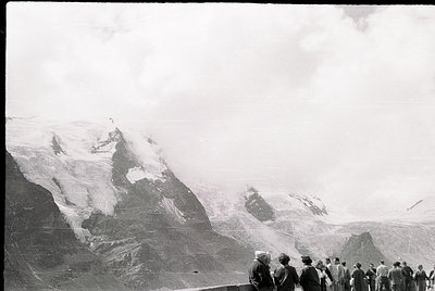 Mid-20th century black-and-white alpine scene: group of hikers in vintage outdoor gear ascends rocky trail toward glacier-cov...