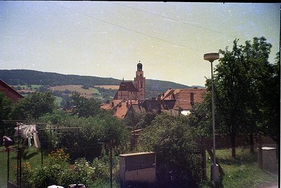 Vintage photograph of a quaint European village with a prominent red-brick church tower dominating the skyline. Foreground fe...