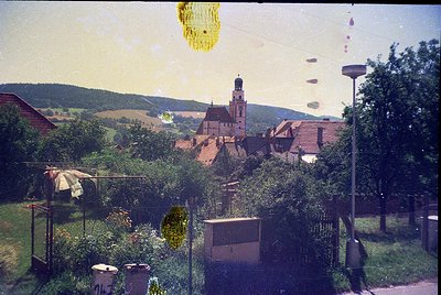 Vintage aerial view of a quaint European village with a prominent castle-like church atop a hill. Lush greenery, rustic homes...