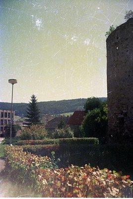 Vintage photo of a rustic European village framed by autumn foliage and a weathered stone tower. Mid-20th century architectur...
