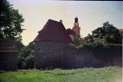Vintage photograph of a weathered stone building with a steep red-tiled roof, surrounded by overgrown greenery. In the backgr...