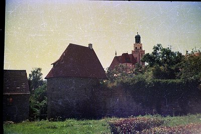 Vintage photo of a rustic stone cottage with a steep red-tiled roof, surrounded by lush greenery and a low hedge. In the back...