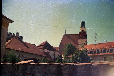 Vintage photograph of European medieval architecture with aged film grain. Red-tiled roofs, a central tower with a bulbous do...