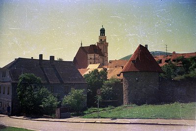 Vintage sepia-toned photo of a European village with a prominent church tower and medieval-style brick tower. Lush greenery s...