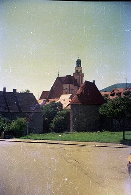 Vintage black-and-white photo of a European church with a steep gable roof and central tower, surrounded by low-pitched resid...