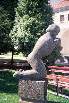 Modernist concrete sculpture of a seated figure in dynamic pose, set on a textured pedestal. Background features lush greener...