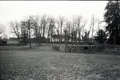 Black-and-white photograph of a barren, overgrown courtyard surrounded by leafless trees and a low stone wall. A single-story...