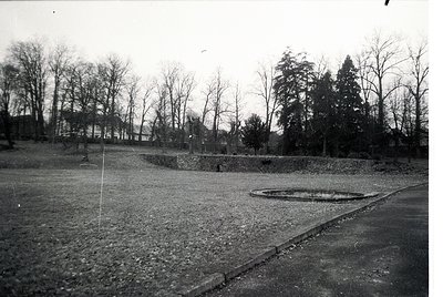Black-and-white photograph of a barren, open courtyard surrounded by bare trees and low stone walls. Circular concrete struct...