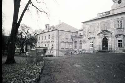 Neoclassical stone building with symmetrical façade, arched doorway, and rectangular windows. Courtyard entrance flanked by b...