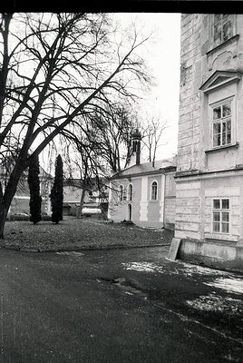 Vintage black-and-white photo of a courtyard featuring a stone church with arched windows and a bell tower, surrounded by lea...