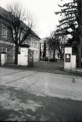 Mid-century European street scene with Soviet-era architecture. Symmetrical gatehouse with "1953" plaque and "1954" sign, fla...