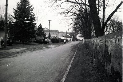 Vintage black-and-white street scene featuring a quiet residential road flanked by a stone wall and sparse trees. Mid-century...