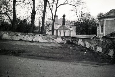 Neoclassical chapel with arched entrance and bell tower framed by leafless trees, set behind weathered stone wall. Likely Eas...