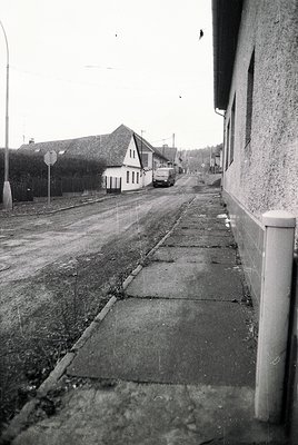 Mid-20th century street scene with cobblestone sidewalk and asphalt road. Whitewashed buildings and a vintage car parked near...