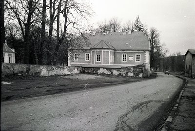 Two-story villa-style building with symmetrical windows, set on a stone foundation. Asphalt road curves left with cobblestone...