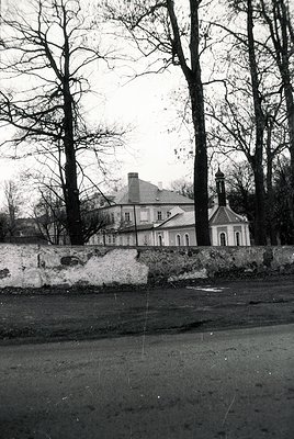 Neoclassical mansion partially obscured by bare winter trees, set behind a stone wall. Symmetrical facade with central chimne...