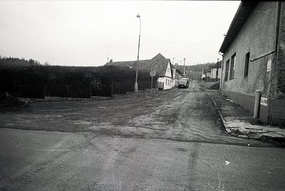 Empty rural street with unpaved sidewalks, lined by utilitarian buildings and sparse vegetation. Mid-century concrete road su...